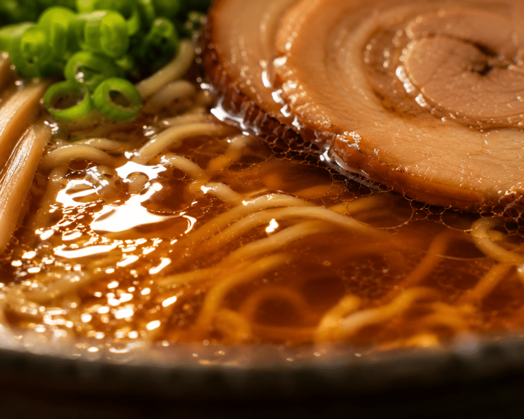 Shoyu ramen: história e características do estilo mais clássico do ramen japonês 2 Close-up do caldo âmbar de shoyu ramen com reflexo de luz, macarrão e chashu ao fundo.